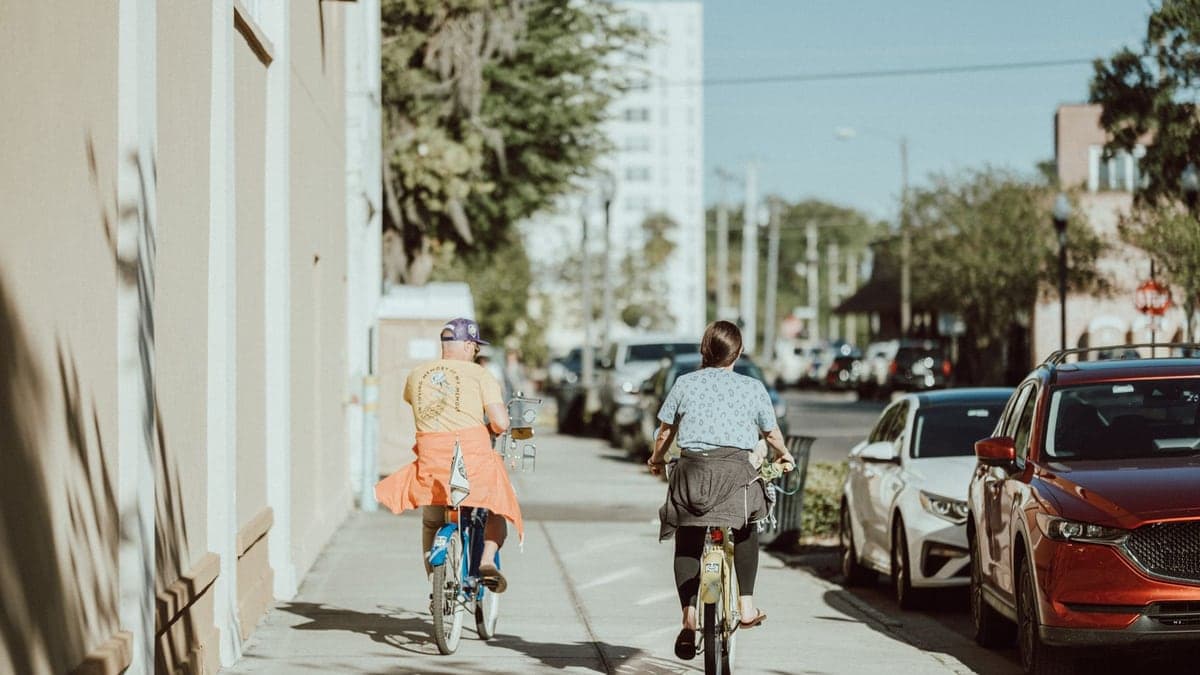 Duas pessoas de costas andando de bicicleta em uma calçada ao lado de carros estacionados em uma rua urbana ensolarada.
