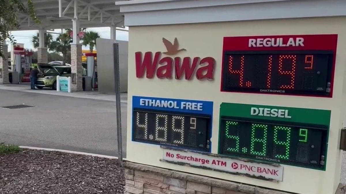 Posto de gasolina com várias bombas e placas de preços sob céu claro em área suburbana com palmeiras e prédios baixos