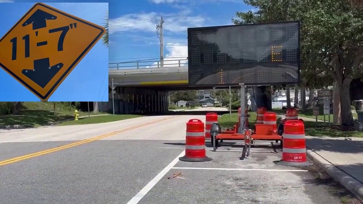 Rua urbana tranquila com ponte ferroviária baixa e placas claras de aviso de altura, pintura brilhante no pavimento, dia ensolarado