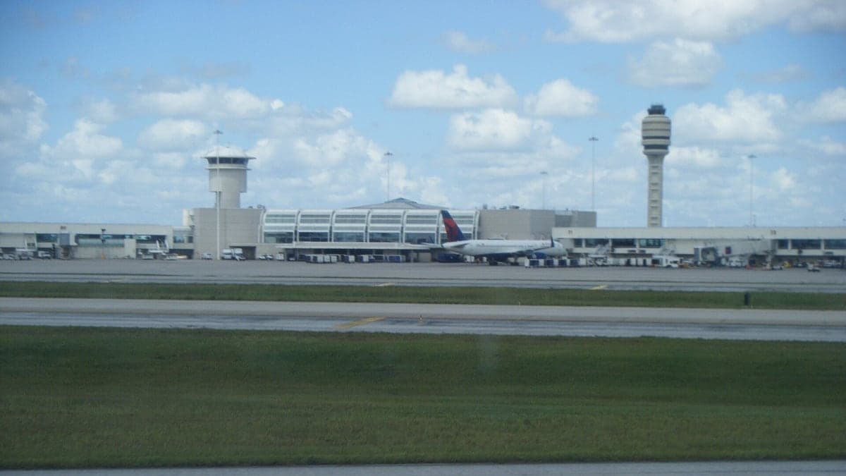 Vista do aeroporto de Orlando com pista, torre de controle e um avião da Delta parado ao lado do terminal sob céu azul com nuvens