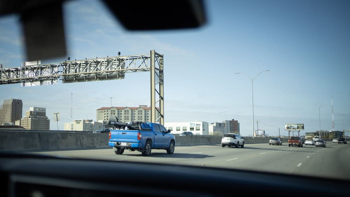 Rodovia interestadual com vários carros trafegando, vista de dentro de um veículo mostrando a pista larga e a cidade ao fundo sob céu azul com poucas nuvens