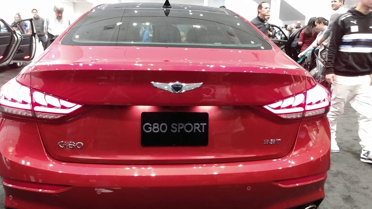 Vista traseira e lateral direita de um carro vermelho modelo G80 Sport da marca Genesis em evento com pessoas ao redor