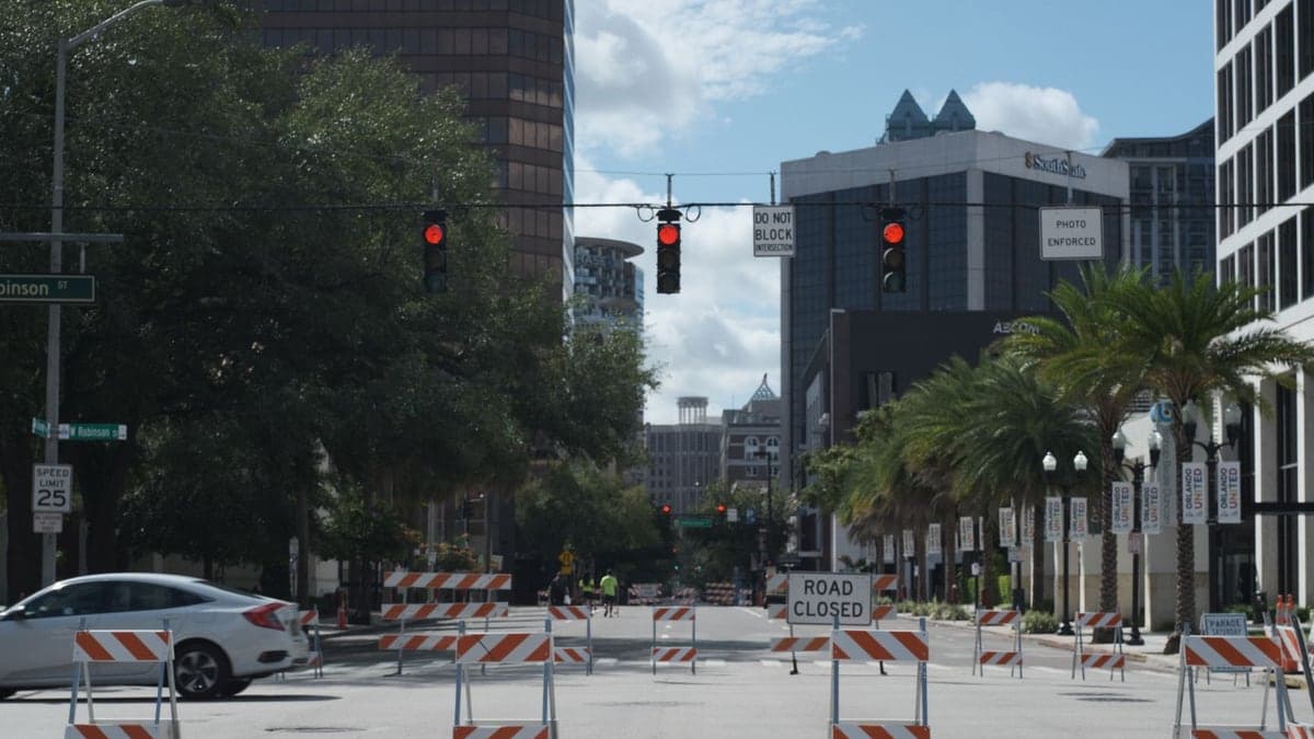 Rua urbana bloqueada com barreiras de construção e sem veículos passando, com semáforos vermelhos ao fundo e prédios ao redor