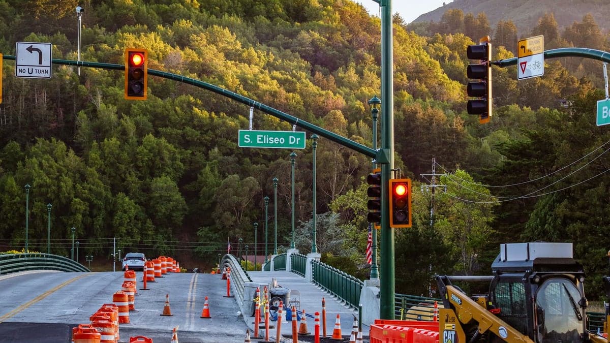 Obras em via urbana com cones laranjas e máquina de construção ao lado de semáforo vermelho em área com vegetação
