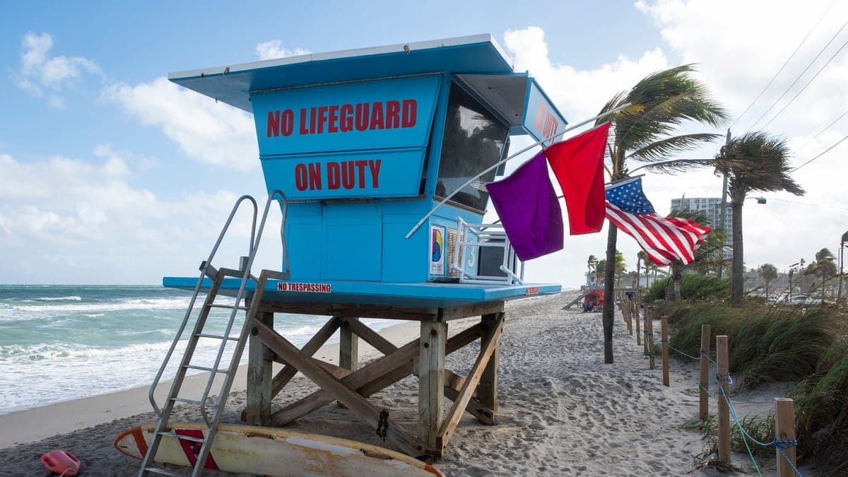 Cabana de salva-vidas azul na praia com placas 'No lifeguard on duty', bandeiras vermelha e roxa ao vento, mar com ondas e céu parcialmente nublado