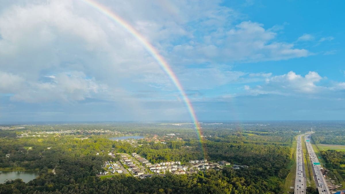 Arco-íris colorido no céu azul com nuvens sobre área verde e linha de casas próximas a uma estrada movimentada em Orlando