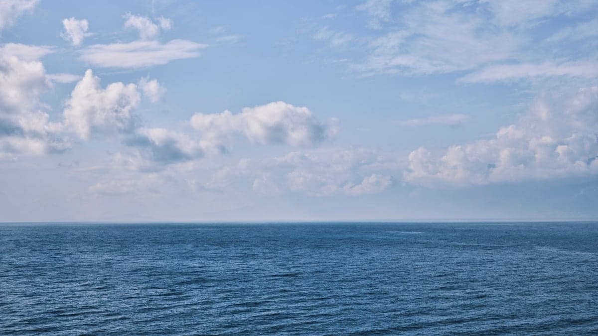 Mar azul sob céu parcialmente nublado com nuvens brancas ao longe, vistos no horizonte.
