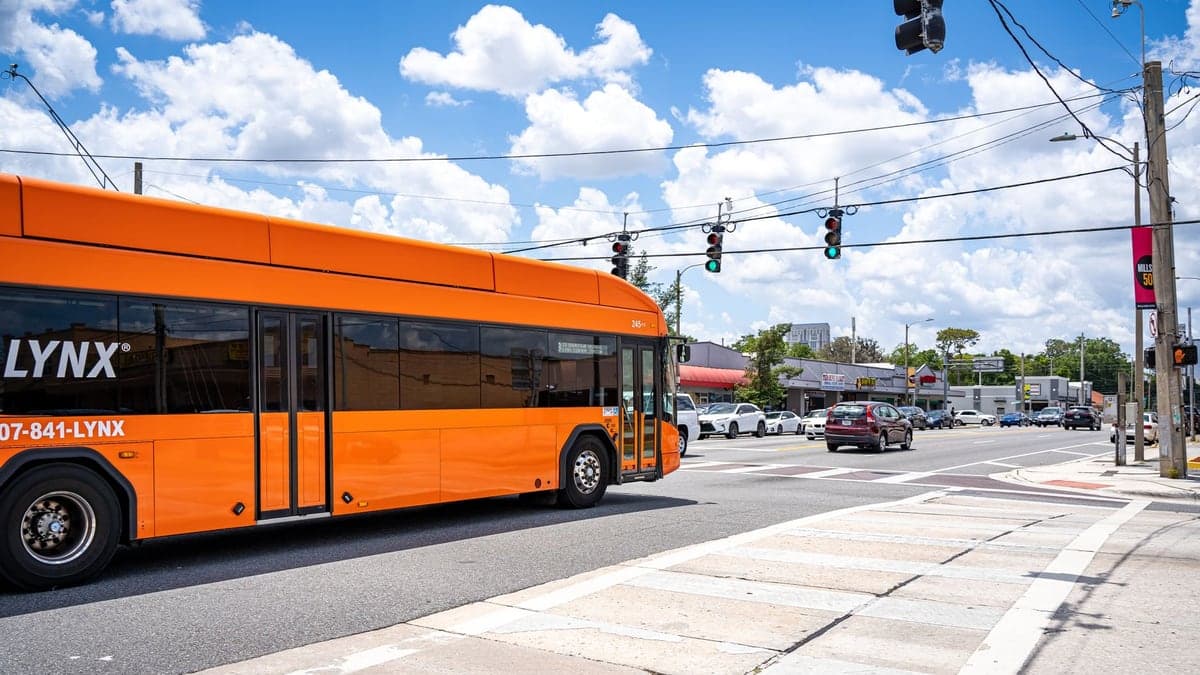 Ônibus laranja da linha LYNX parado em semáforo em uma avenida movimentada sob céu azul com nuvens