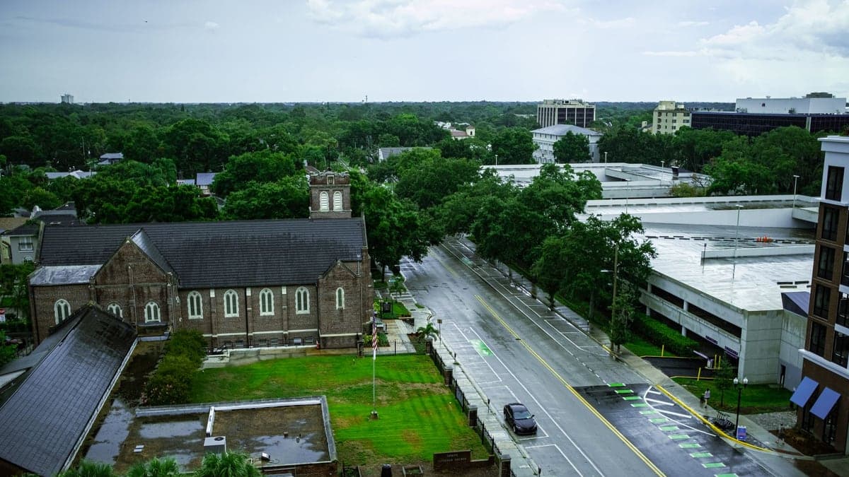 Vista aérea de rua em Orlando com céu parcialmente nublado e chão úmido, mostrando árvore, igreja e prédios ao redor