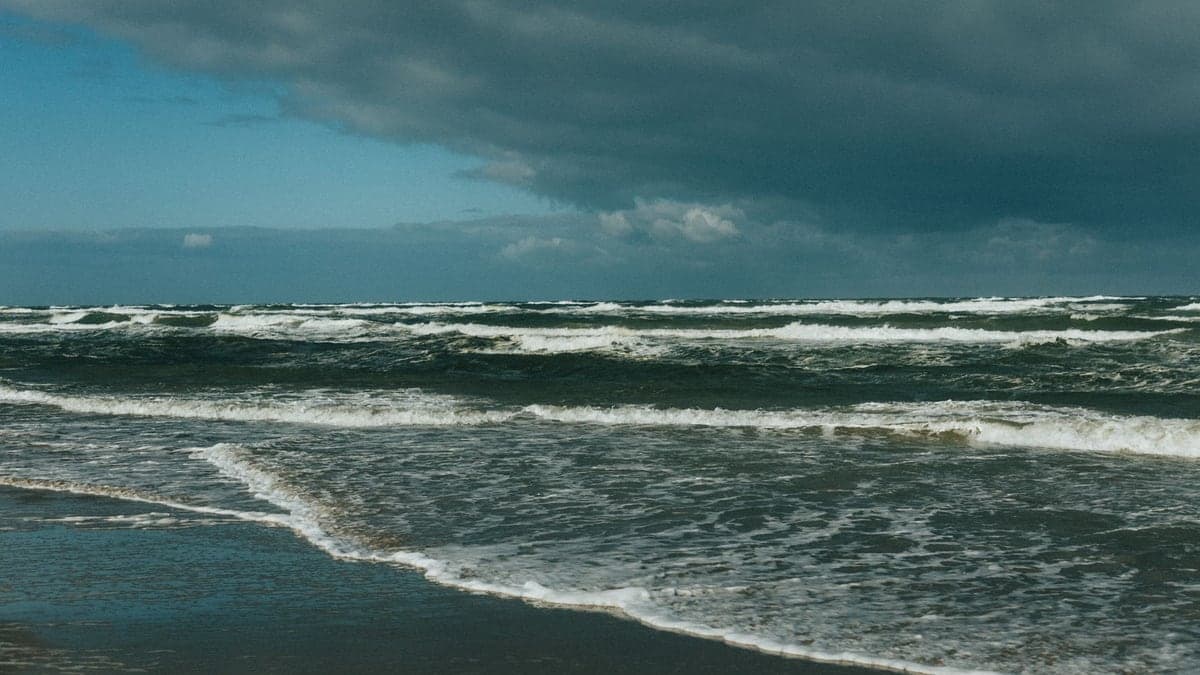 Praia com mar agitado, ondas grandes e céu parcialmente nublado com nuvens escuras ao fundo
