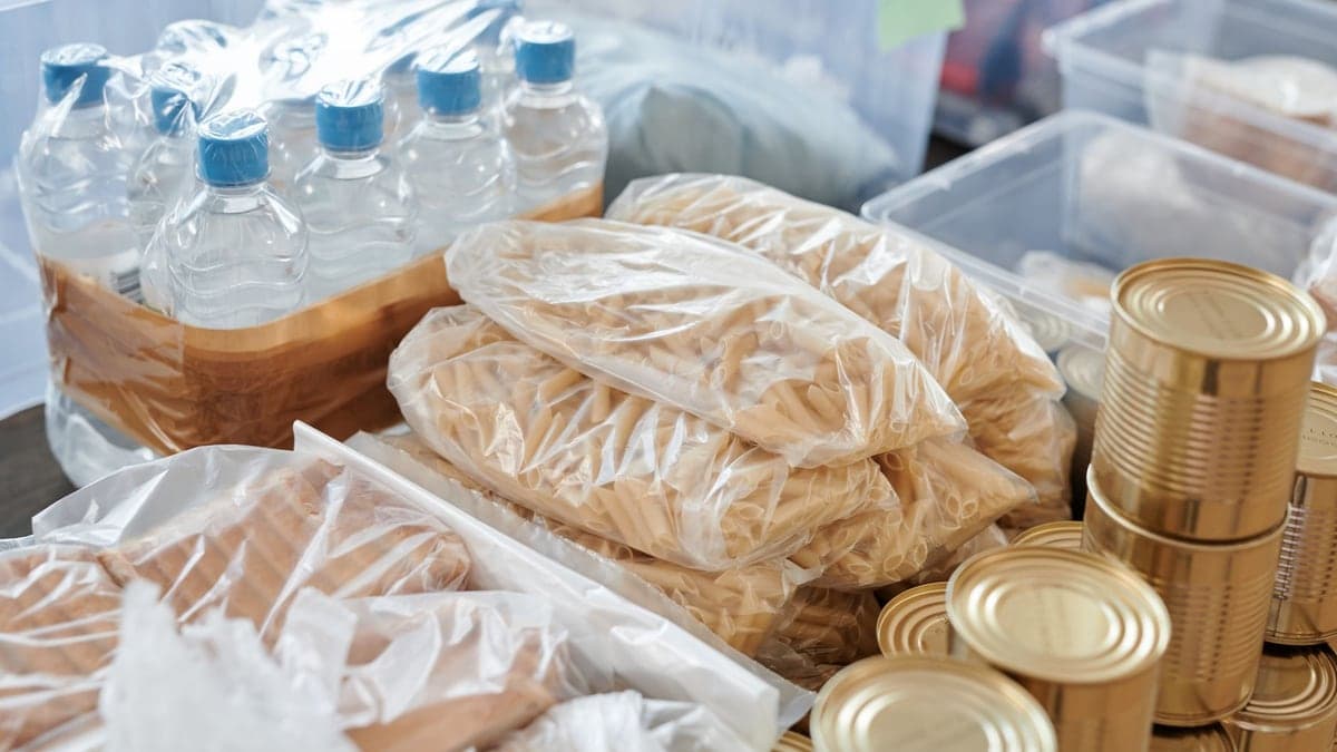 mesa com alimentos embalados, incluindo garrafas de água, pacotes de macarrão e latas, organizados para distribuição
