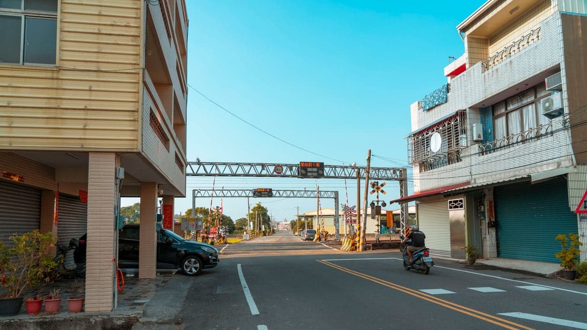 Cruzamento de trem em área urbana com sinais de trânsito e equipamentos ferroviários ao longo da via, ao fundo postes e veículos estacionados