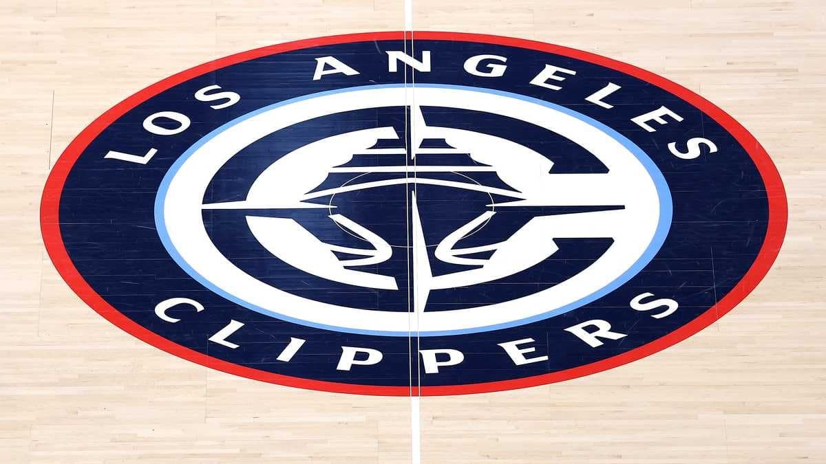 Fachada externa da arena da NBA, com estacionamento e palmeiras ao redor, sob céu azul
