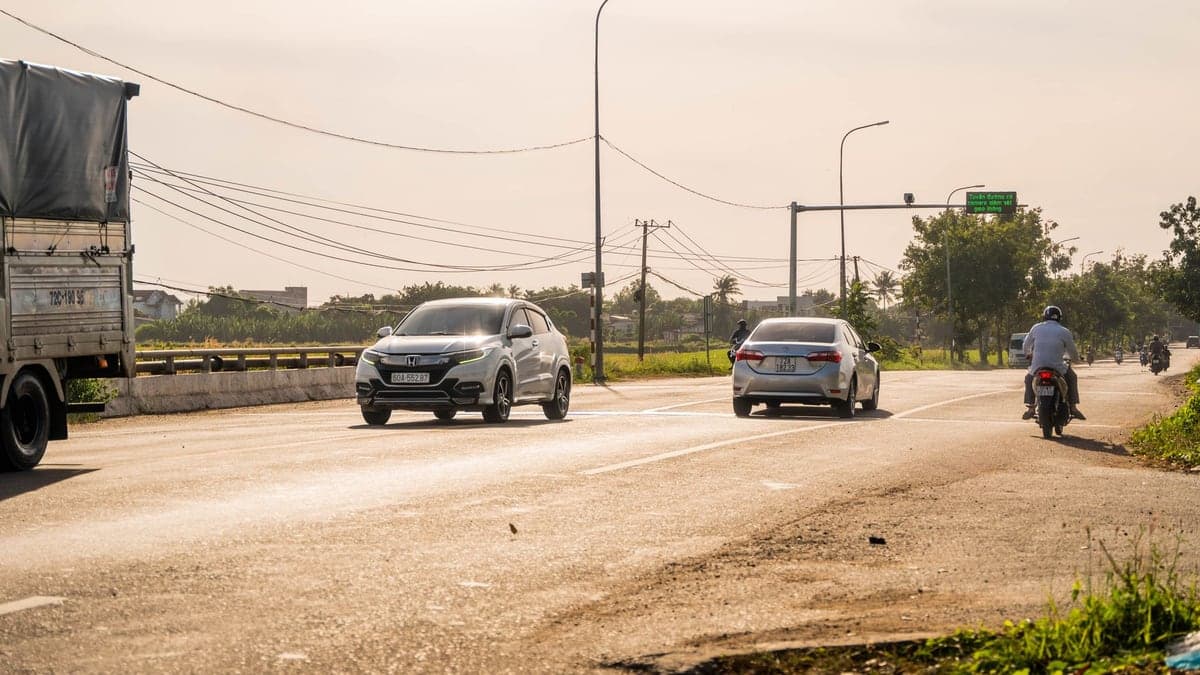 Rodovia reta com poucos veículos, incluindo dois carros e uma motocicleta, em cenário de fim de tarde com poste de iluminação e vegetação ao lado da estrada