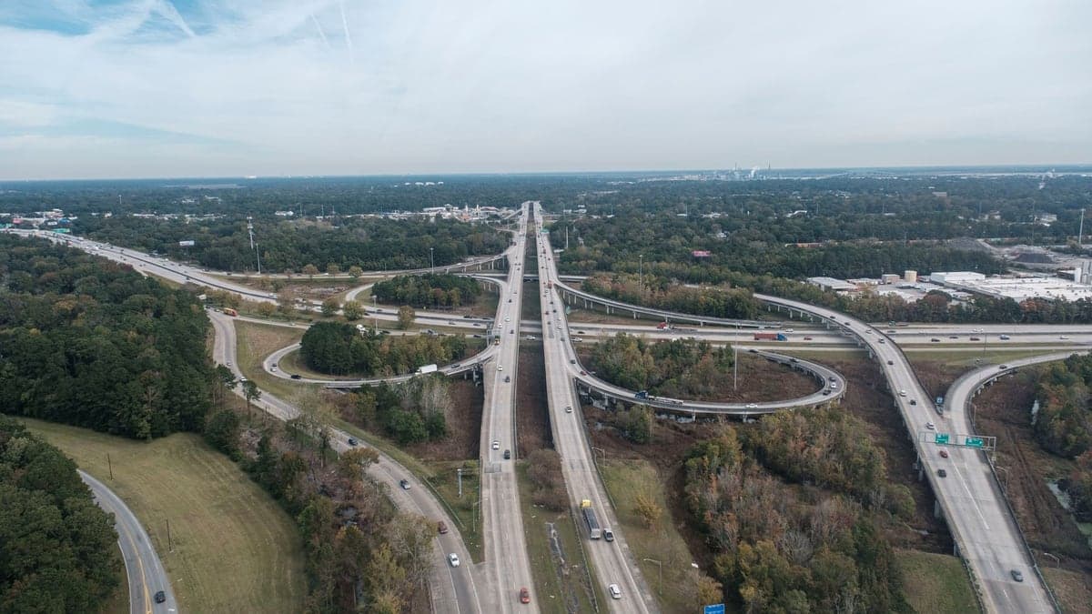 Vista aérea diurna de uma rodovia com várias faixas e viadutos, cercada por áreas verdes e algumas construções ao fundo