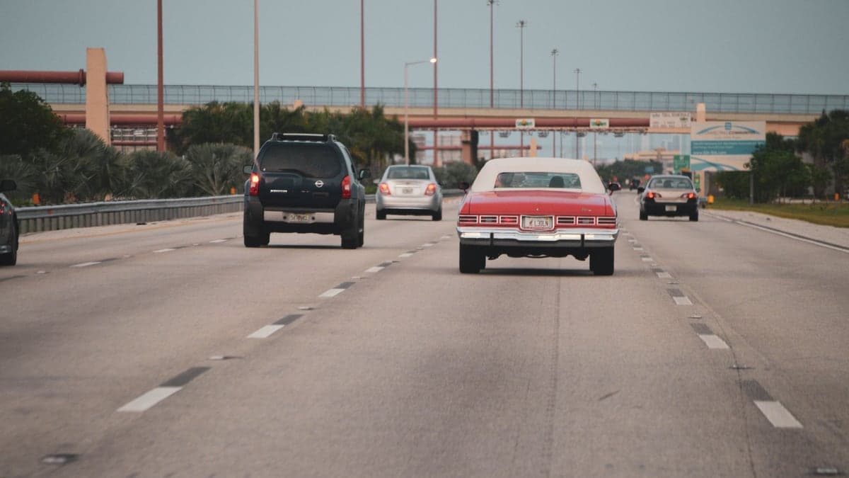Imagem de uma via expressa movimentada com vários carros, faixa larga e viaduto ao fundo, em um ambiente urbano.