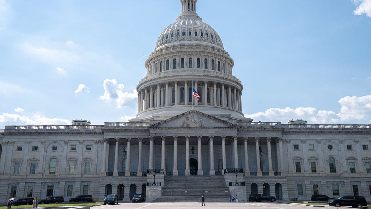 Fachada do Capitólio dos Estados Unidos com a cúpula branca e uma bandeira americana ao centro, sob céu azul com algumas nuvens.