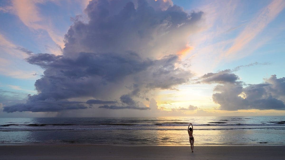Praia com mar calmo no pôr do sol, grande nuvem escura no céu e uma pessoa em pé na areia de costas com os braços levantados