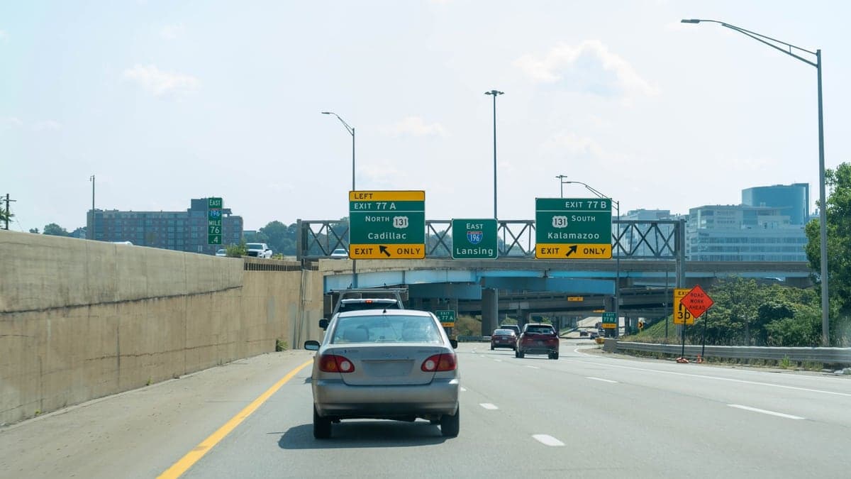Carro dirigindo em rodovia com semáforo e placas de saída, com edifícios ao fundo em área urbana