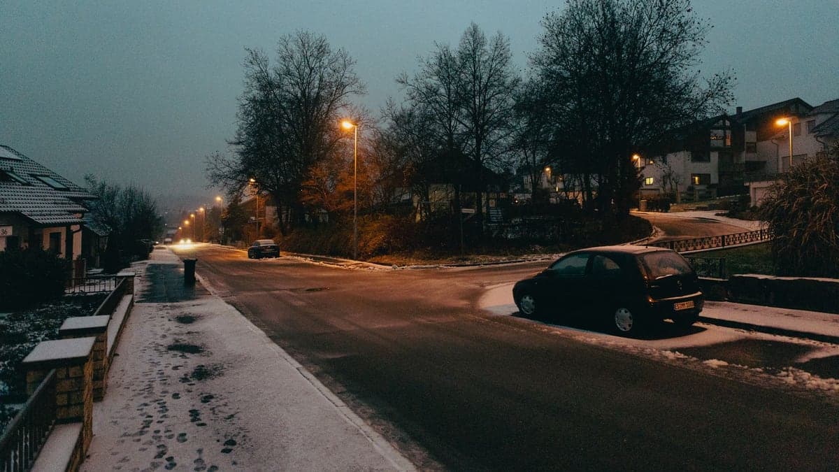 Rua residencial durante o entardecer com postes de luz acesos próximos a casas, uma árvore e um carro estacionado perto de uma rotatória ao fundo.