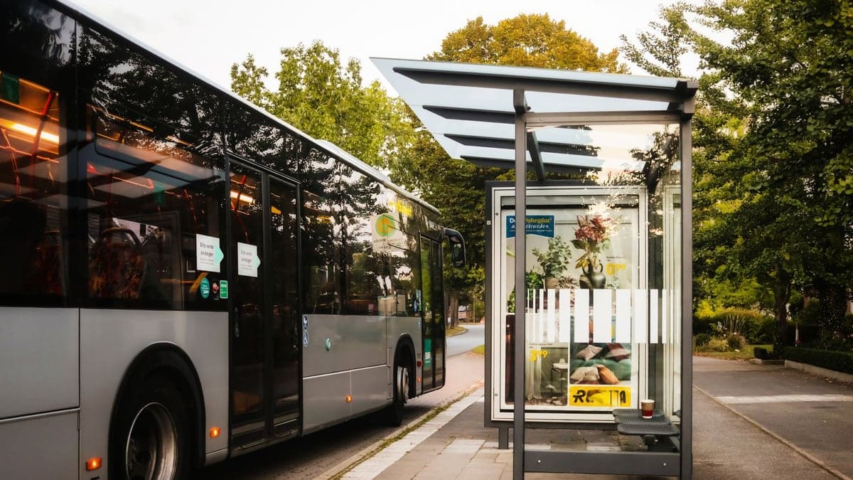 ônibus preto parado ao lado de ponto de ônibus com abrigo de vidro em rua cercada por árvores