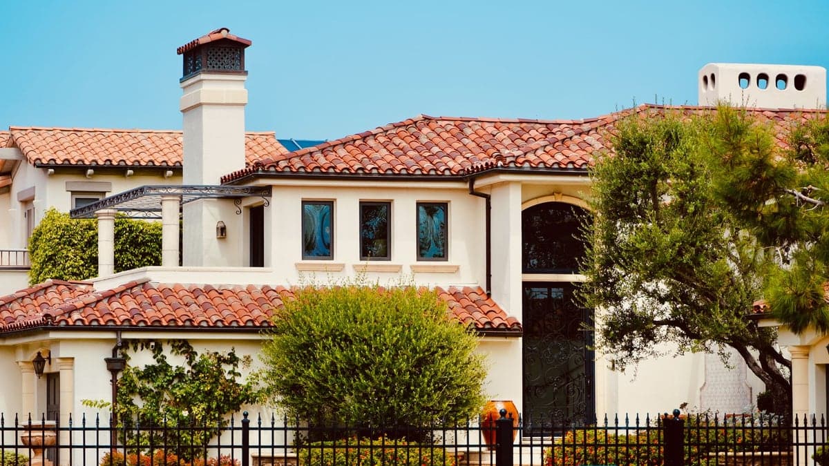 casa com telhado de telhas vermelhas, jardim na frente e cerca preta, vista lateral com árvores e céu azul