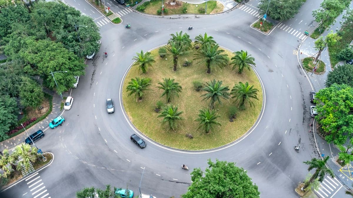 Vista aérea de um cruzamento em rotatória com várias faixas de pedestres e carros circulando, rodeado por áreas verdes e árvores.